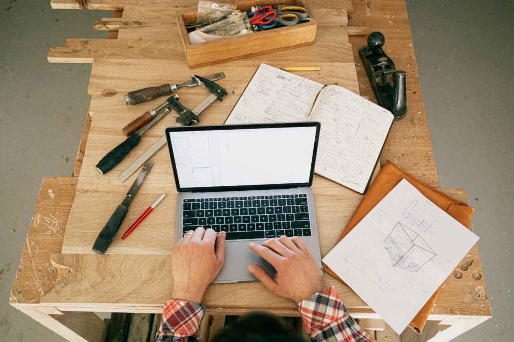 Carpenter using laptop for woodwork designs at a workbench in a workshop.